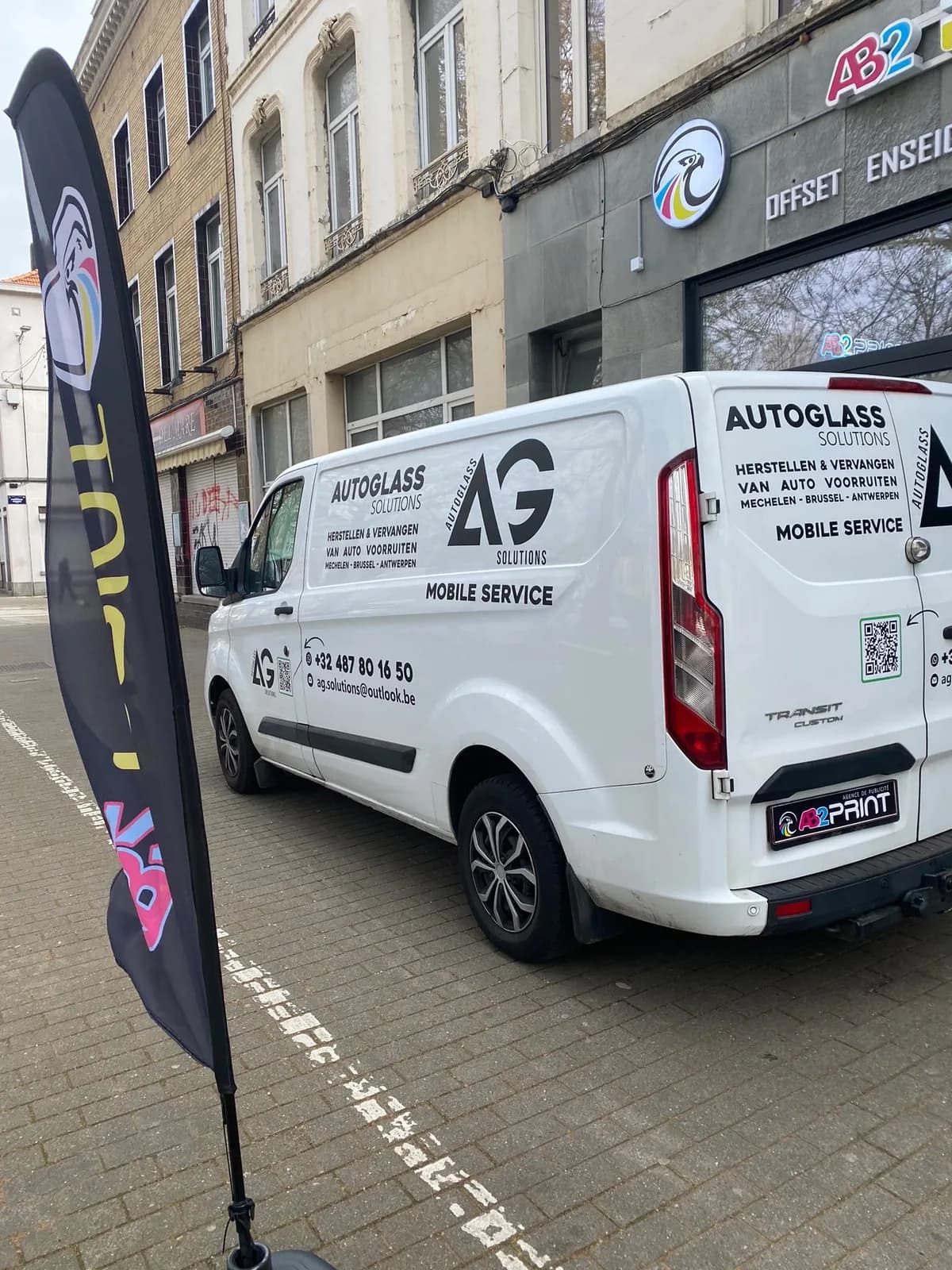 Façade de l'atelier AB2 Print à Molenbeek avec son enseigne, une camionnette habillée et un beach flag devant la vitrine